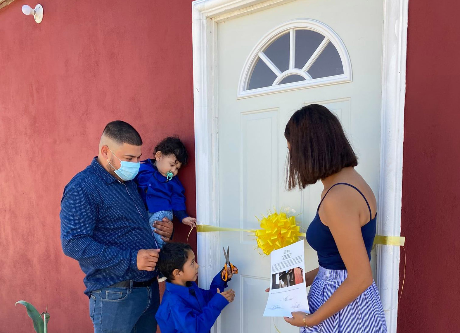 Family with 2 little kids cut the ribbon to their newly completed home, painted red