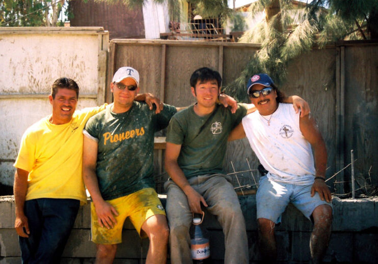 4 men sit arm in arm smiling at a worksite, including Steve a white man, Eduardo a mexican man, and two others